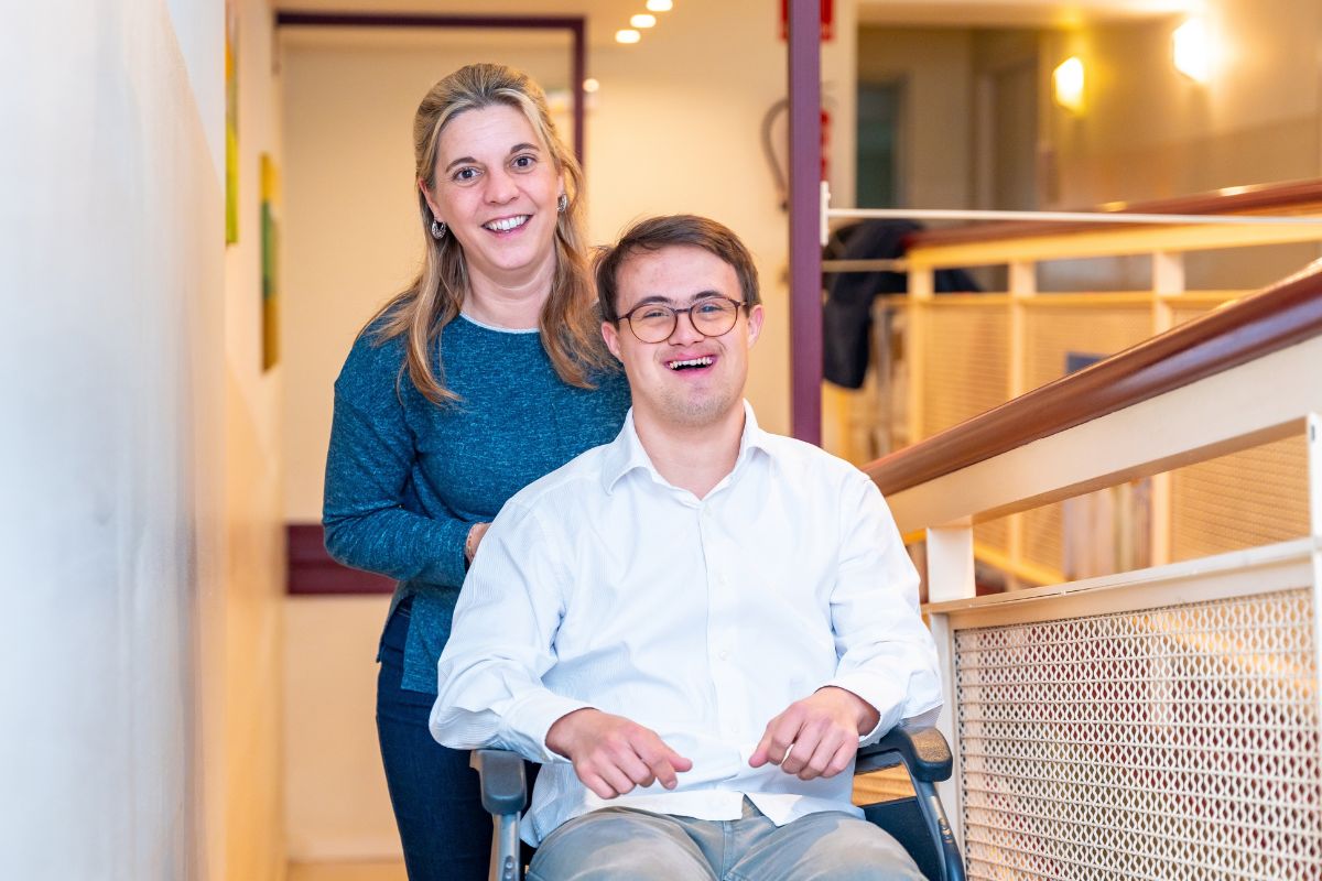 A smiling young man being pushed in a wheelchair by a blonde woman assumed to be his carer.